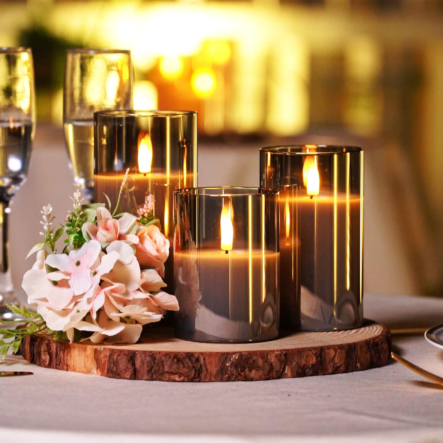 Fitlybuy AuraFlame LED candles in smoke-grey glass cylinders on a wooden base with pink flowers and warm lighting.