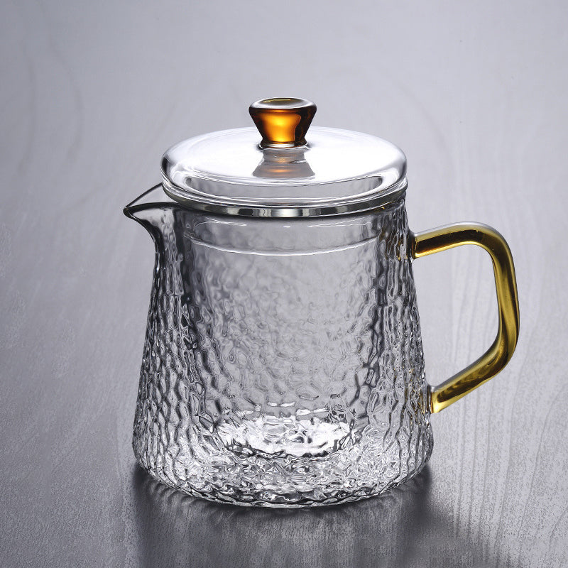 Clear, heat-resistant glass teapot with a hammered texture, golden handle, and amber knob on lid against a gray background.