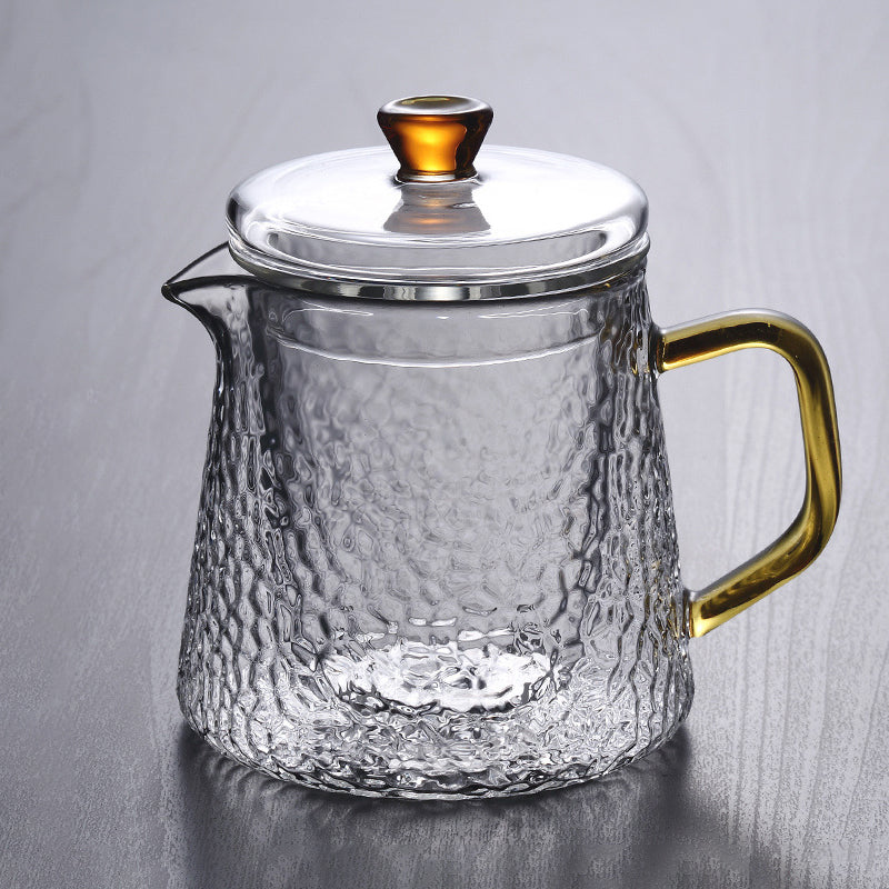Clear, hammered glass teapot with a golden handle and amber-toned knob on a gray background.