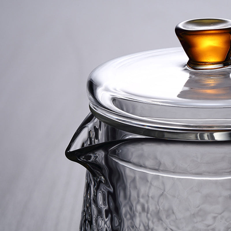 Close-up of a clear, textured glass Pindefang teapot with a glowing amber lid knob against a light gray background.
