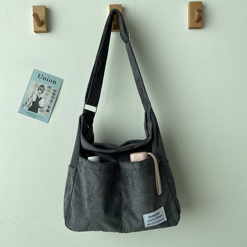 Grey corduroy crossbody bag with front pockets and adjustable strap, hanging on a wall hook next to a wall card.