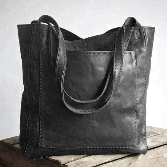 Fitlybuy Eva black leather tote bag with a large outer pocket and shoulder straps, resting on a rustic wooden surface.