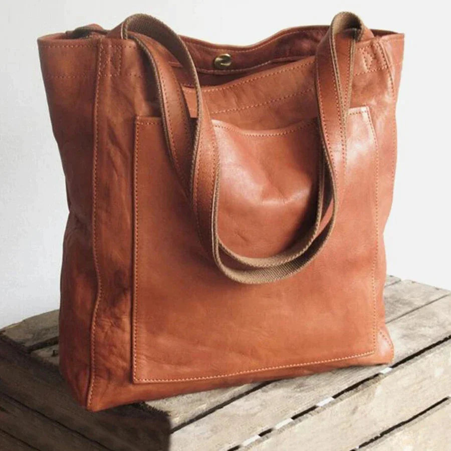Tan leather tote bag with dual shoulder straps and a front pocket, sitting on a wooden crate.