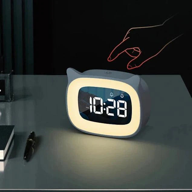 Gray cat-eared LED alarm clock with glowing night light and digital display, sitting on a desk with a hand touching top.