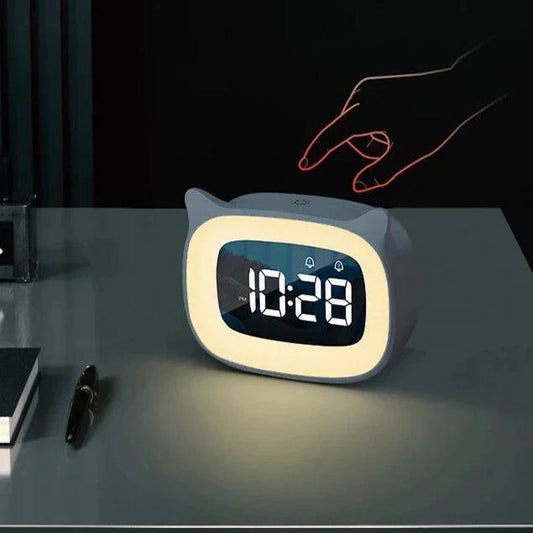 Gray cat-eared LED alarm clock with glowing night light and digital display, sitting on a desk with a hand touching top.