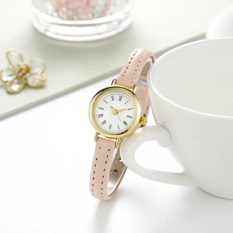 Fitlybuy Billie gold-tone round watch with a white Roman numeral dial and blush pink leather strap on a desk.
