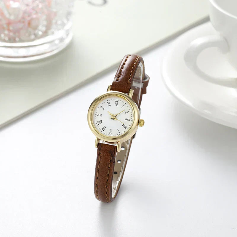 Fitlybuy Billie minimalist gold watch with white dial, Roman numerals, and brown leather strap on a white table.