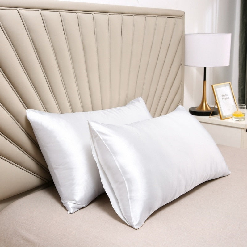 Two white Fitlybuy satin pillowcases on a bed with a beige tufted headboard and bedside lamp.