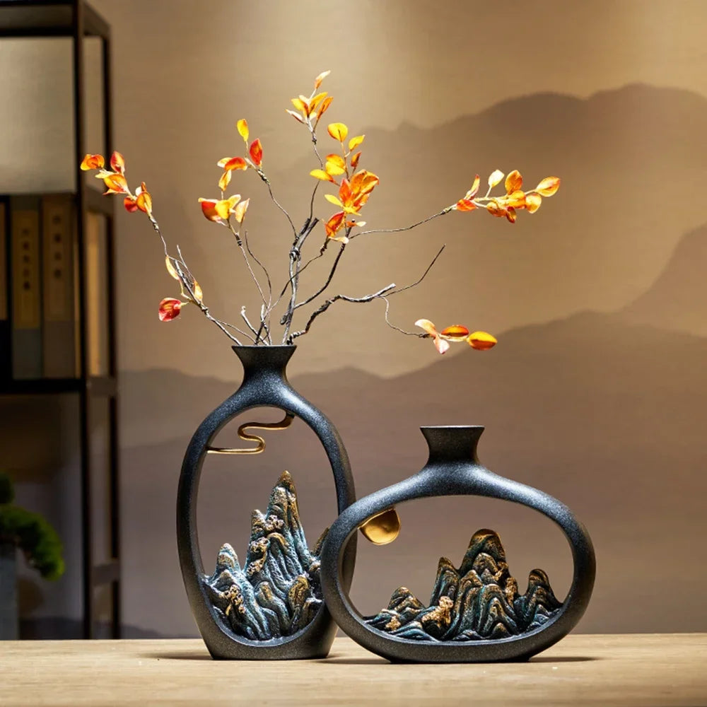 Two black ceramic vases with mountain landscape cutouts and gold accents, displayed on a table with orange leaf stems.