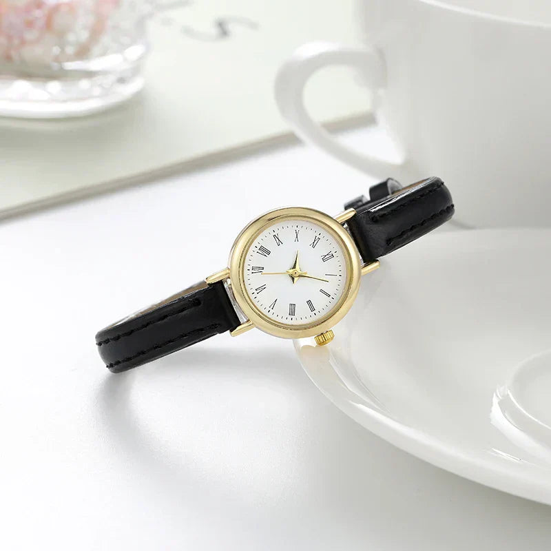 Fitlybuy gold round women’s watch with a white dial, Roman numerals, and a black leather strap on a white table.