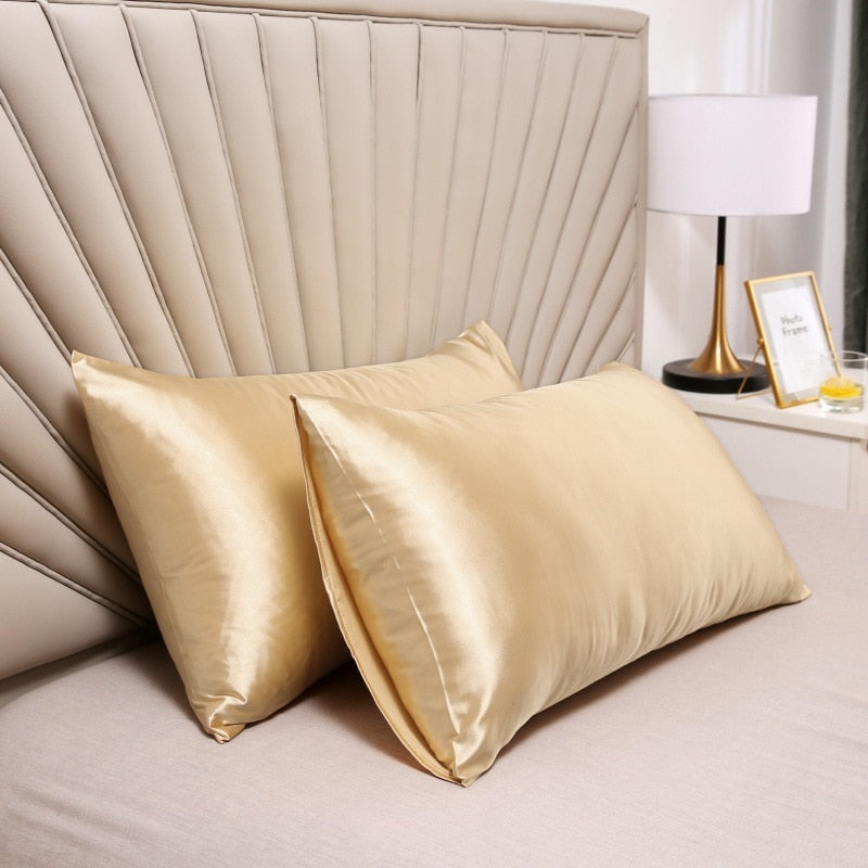 Two shiny gold Fitlybuy satin pillowcases on a bed with a beige headboard, lamp, and side table in the background.