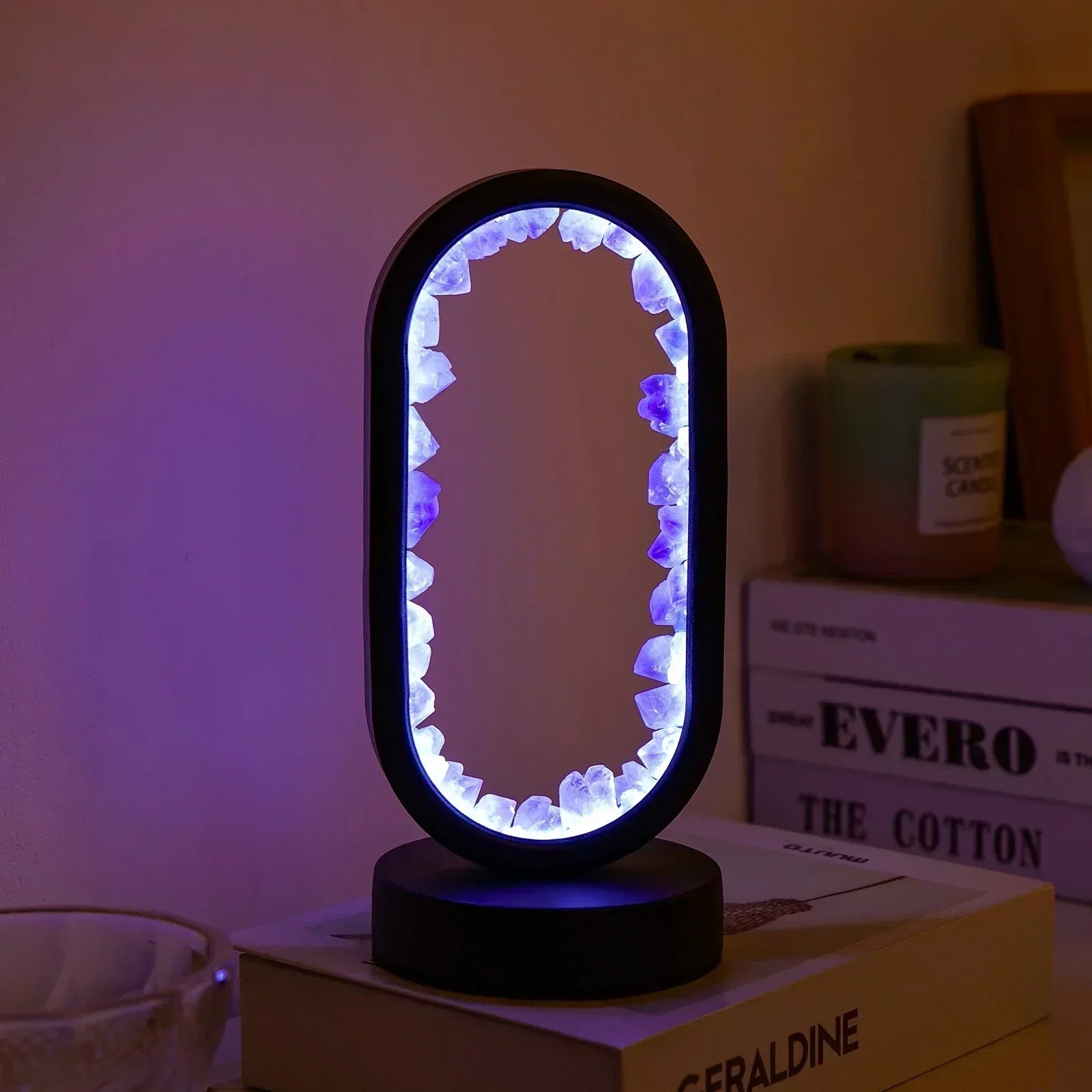 Oval Fitlybuy AuraStone lamp with glowing amethyst crystals inside a black frame on a stack of books in a cozy room.