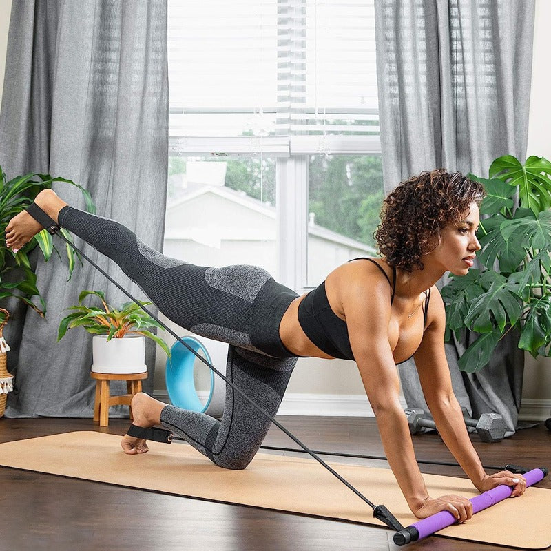 Woman using purple Fitlybuy Pilates bar with resistance bands for leg extension exercise on a yoga mat at home.