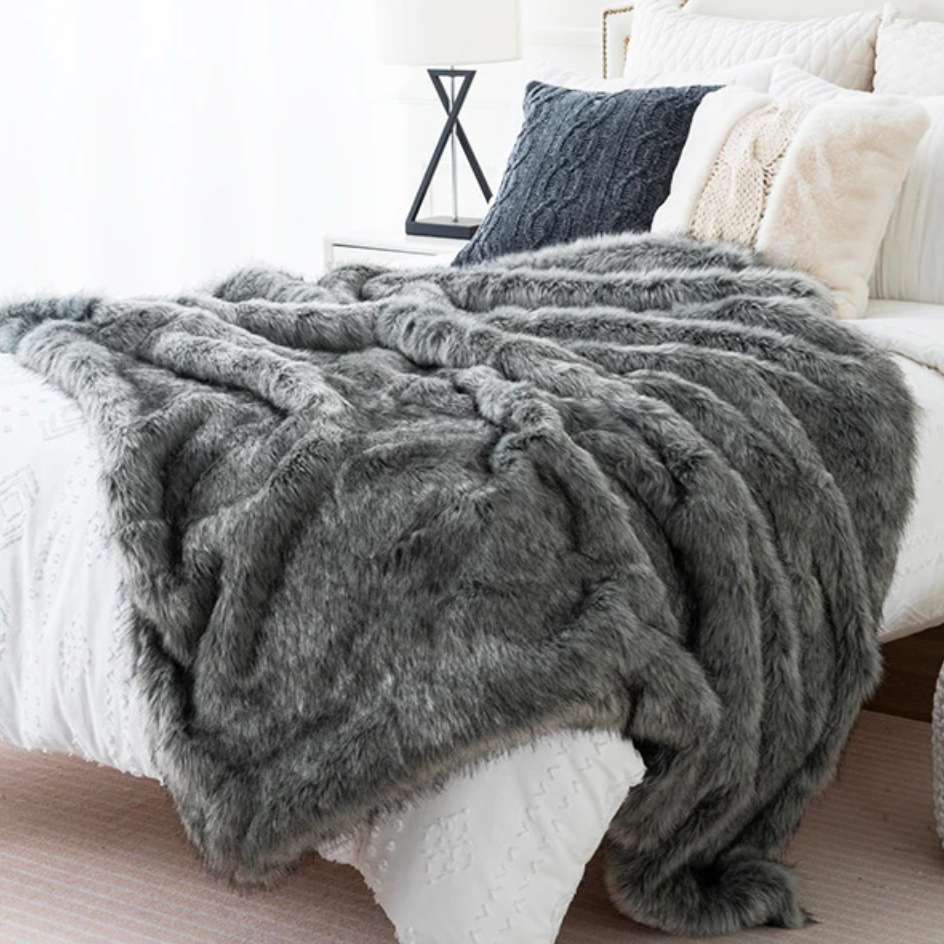 Grey faux fox fur throw blanket draped over a white bed with decorative pillows in a bedroom setting.