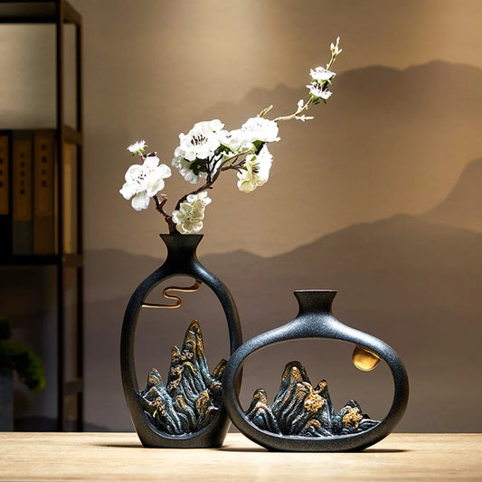 Two black ceramic vases with gold-accented mountain landscape cutouts, one holding a branch of white faux flowers.