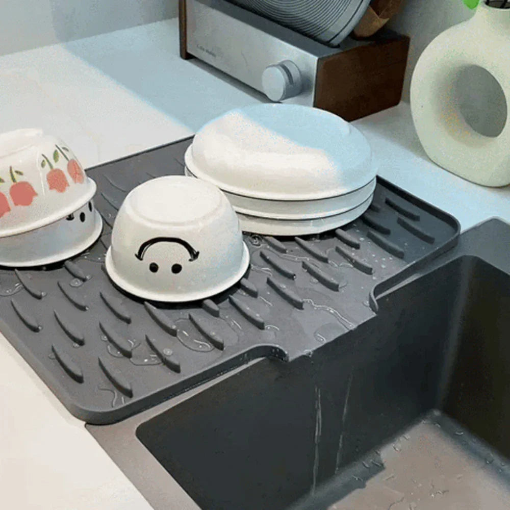 Gray Fitlybuy Silicool silicone drying mat with raised ridges, draining water into a kitchen sink with bowls on top.
