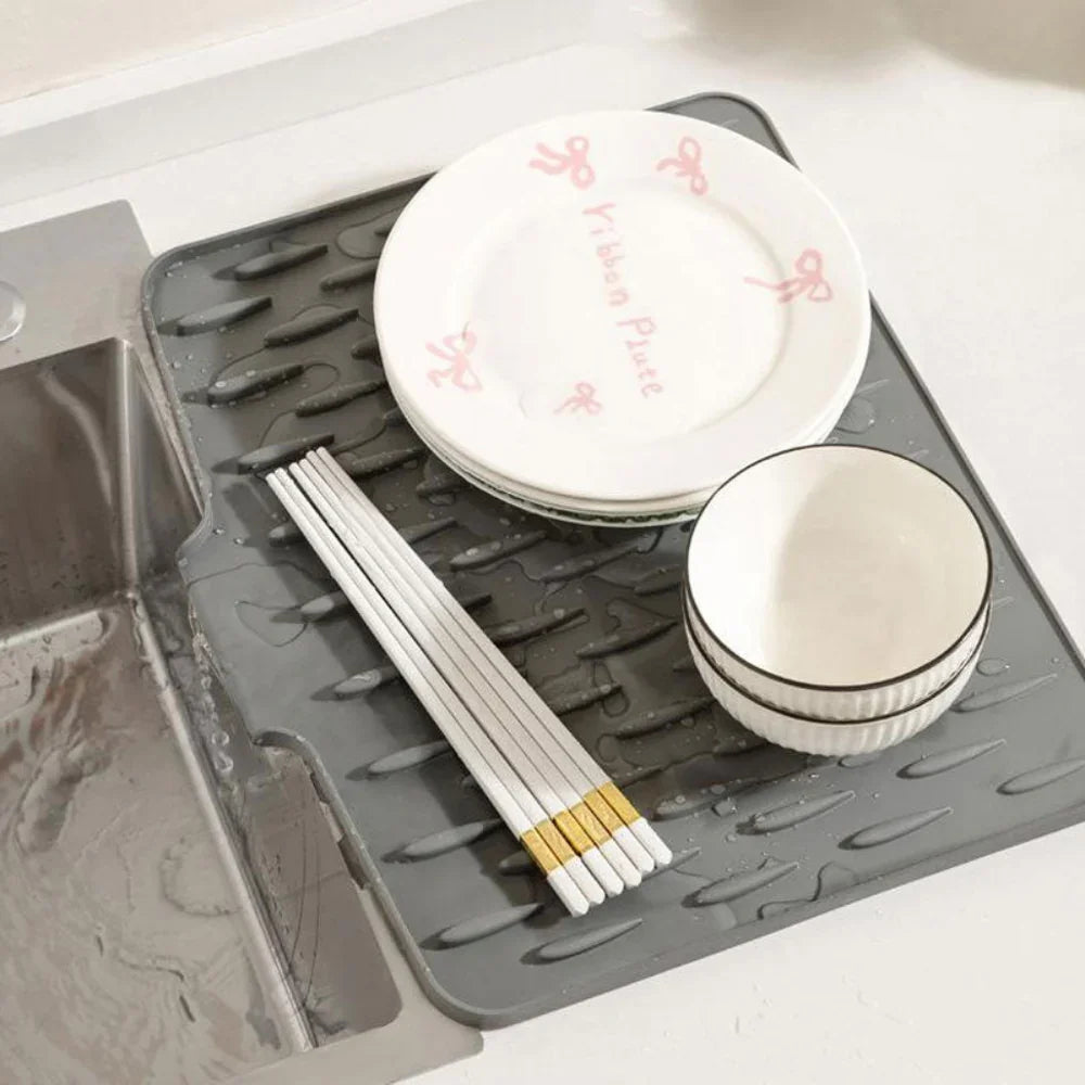 Grey silicone dish drying mat with ridges holding plates, bowls, and chopsticks next to a kitchen sink.