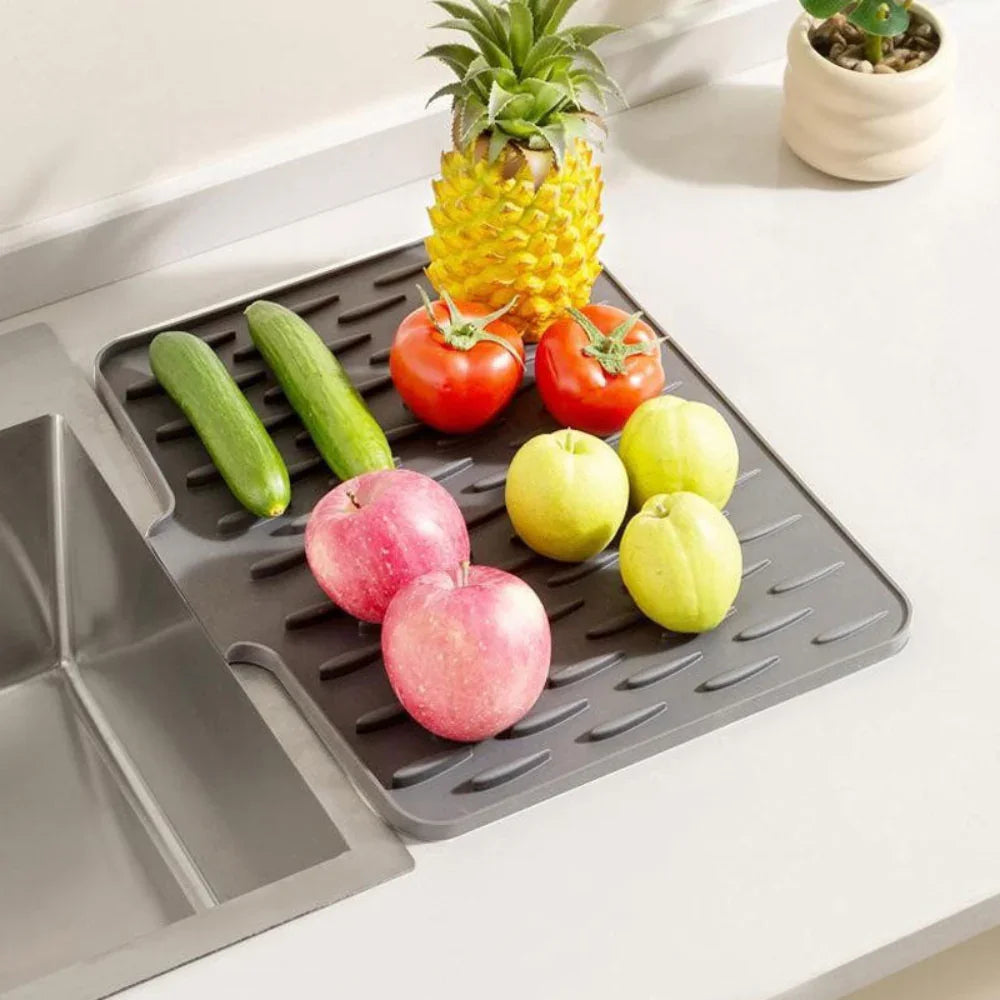 Gray, non-slip silicone drying mat next to a kitchen sink, holding fresh fruits and vegetables.
