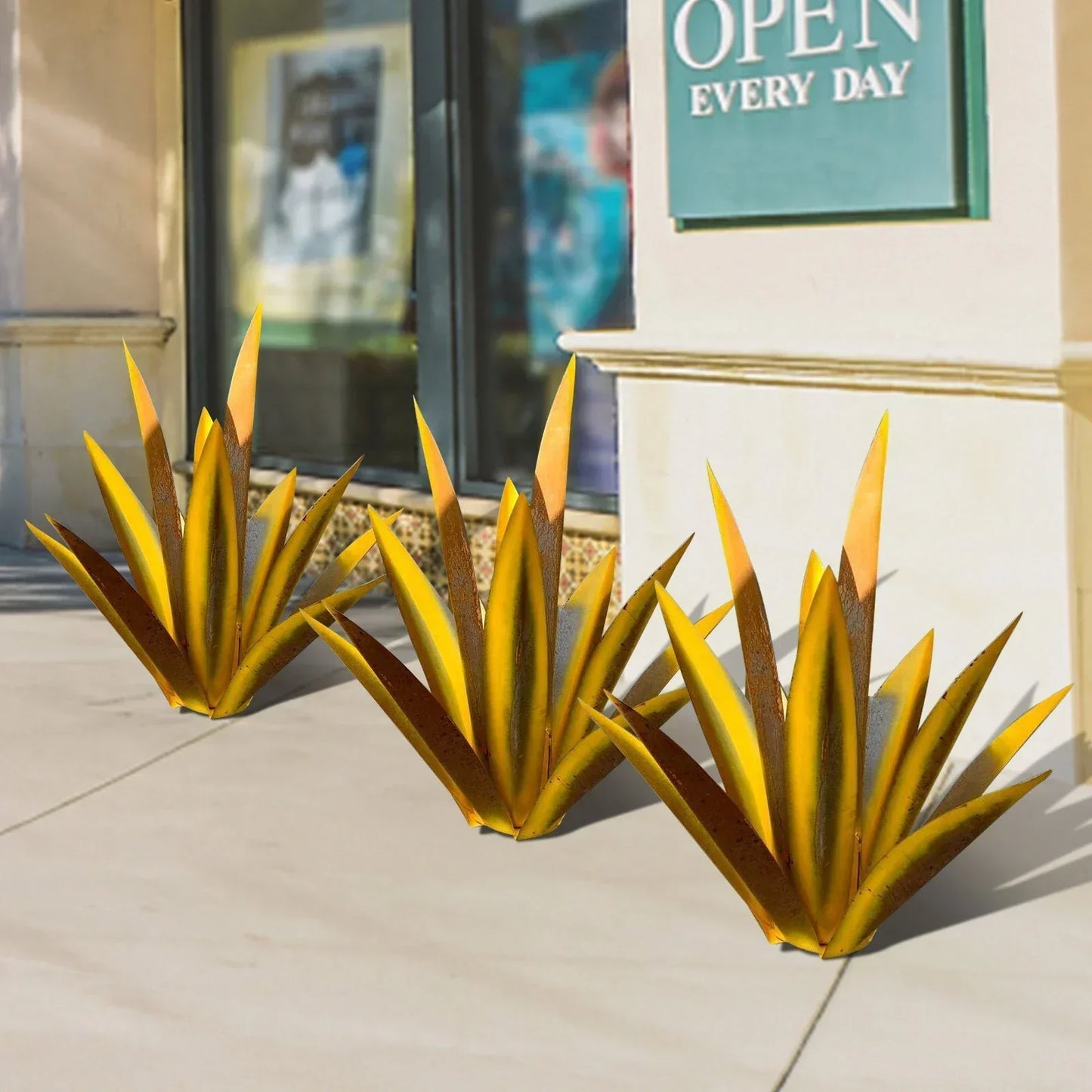 Three handcrafted yellow and rust-finished metal agave sculptures displayed on a concrete sidewalk.