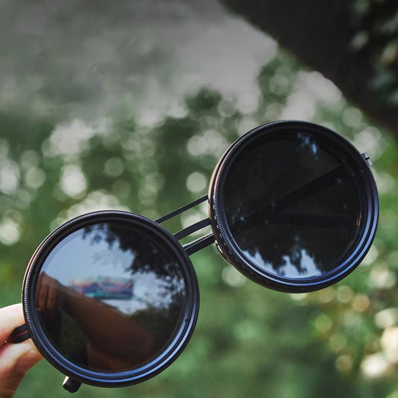 Fitlybuy unisex round polarized sunglasses with black metal frames held against a blurred outdoor nature background.