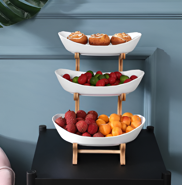 Three-tiered Fitlybuy fruit bowl with white ceramic trays and a light wood frame, holding snacks and fresh fruit.