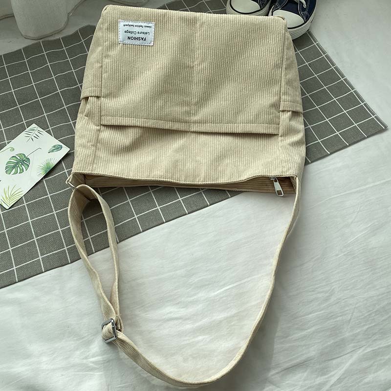 Beige corduroy crossbody bag with a front pocket and label, on a plaid mat with white fabric background.