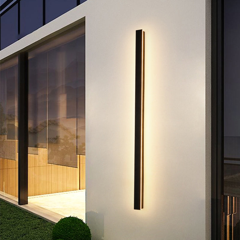Fitlybuy modern black minimalist LED wall light mounted vertically on an outdoor white wall, emitting a warm glow.