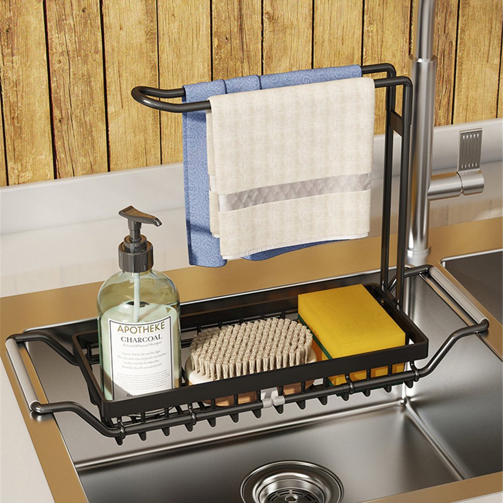 Black adjustable over-sink organizer rack holding a soap bottle, brush, and dish towels in a stainless steel kitchen sink.