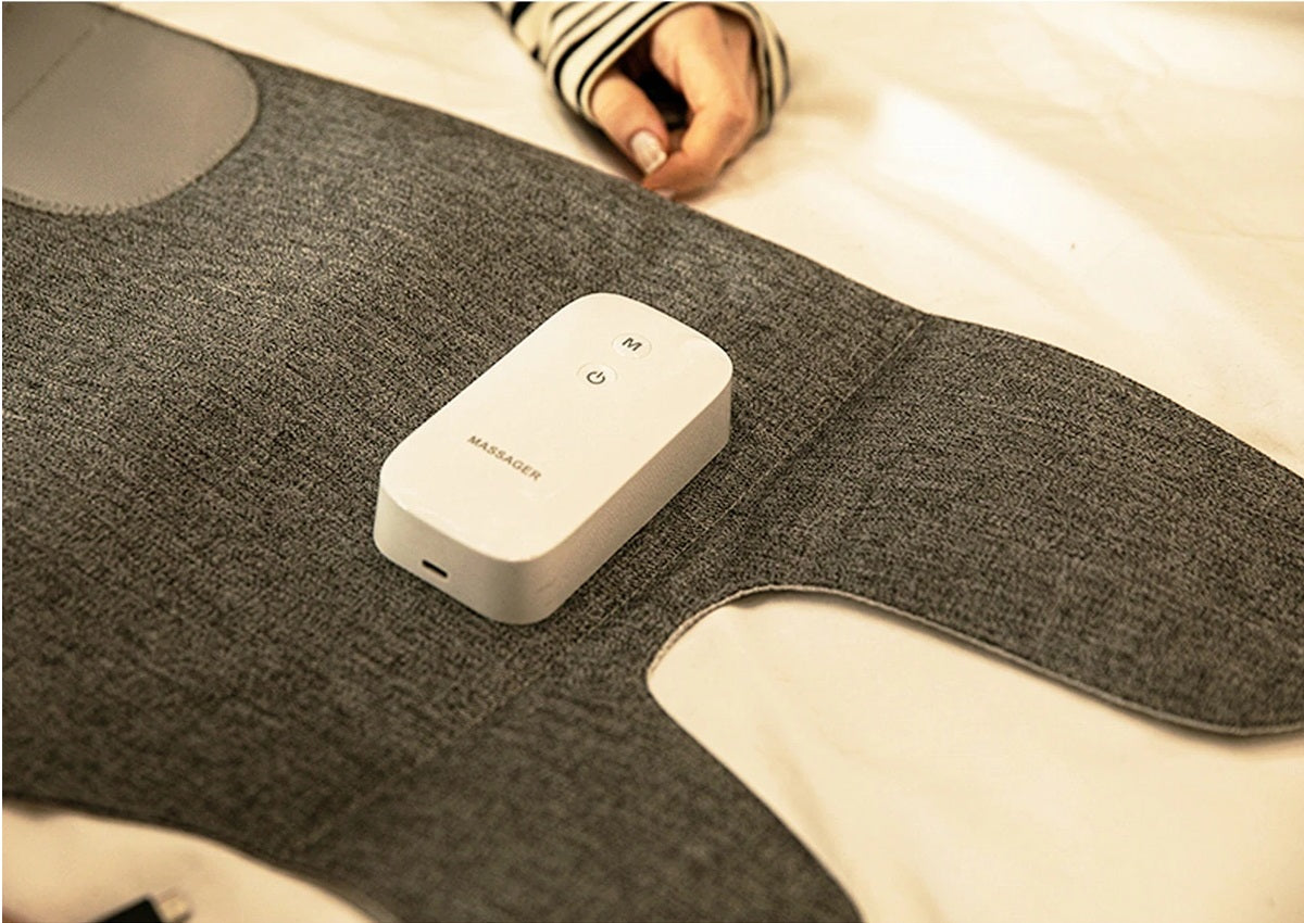 Fitlybuy grey fabric leg massager with a white handheld controller on a bed, showing mode and power buttons.