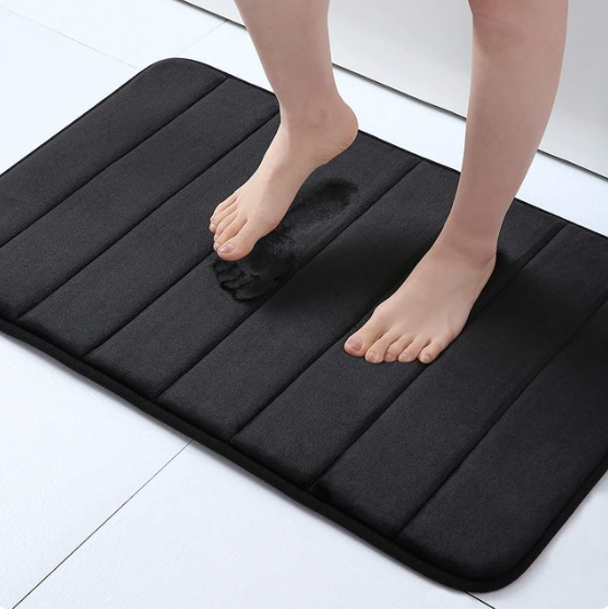 Person standing on a black, striped Fitlybuy memory foam bath mat showing a footprint indentation for absorption.
