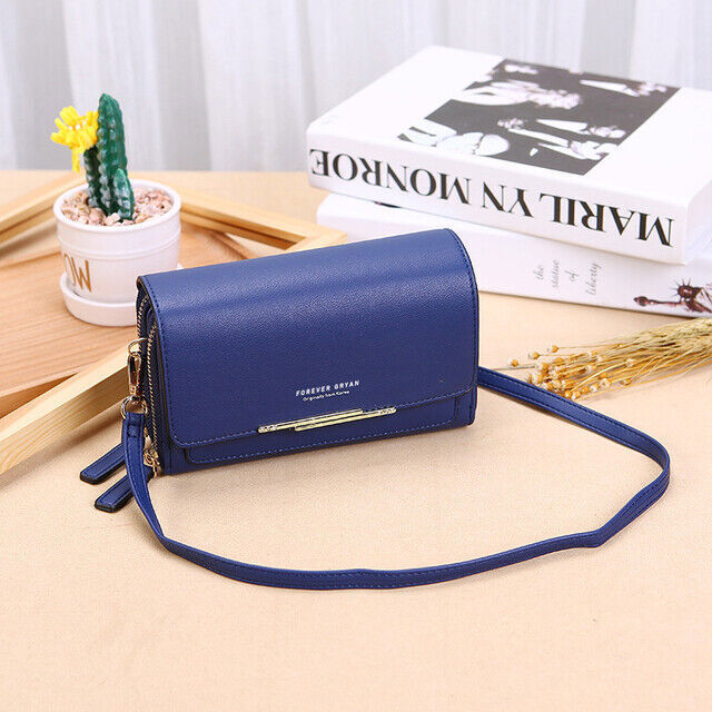 Blue leather crossbody bag with gold accent, zippers, and strap, displayed on a table near books and a small plant.
