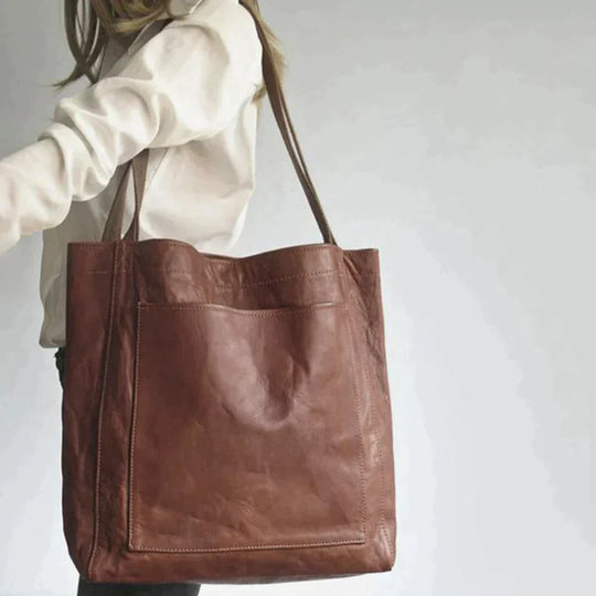 Woman carrying a brown, spacious leather tote handbag with a minimalist design and large front exterior pocket.
