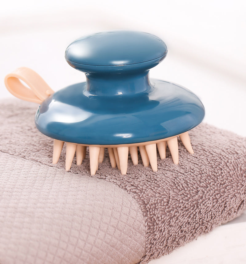 Blue Fitlybuy Scalp Bliss Pro massager with beige silicone bristles and ergonomic handle, resting on a brown towel.