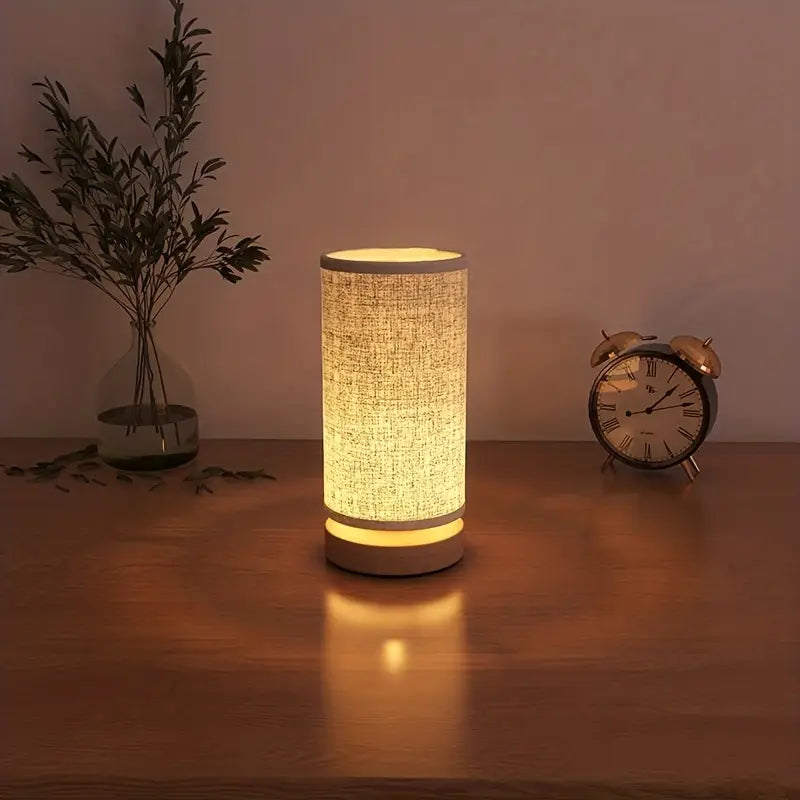 Fitlybuy minimalist cylindrical wooden lamp with fabric shade glowing with soft warm light on a wooden bedside table.