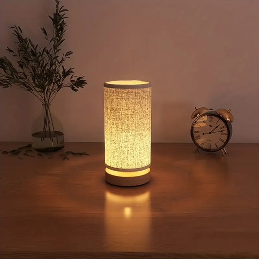 Fitlybuy minimalist cylindrical wooden lamp with fabric shade glowing with soft warm light on a wooden bedside table.