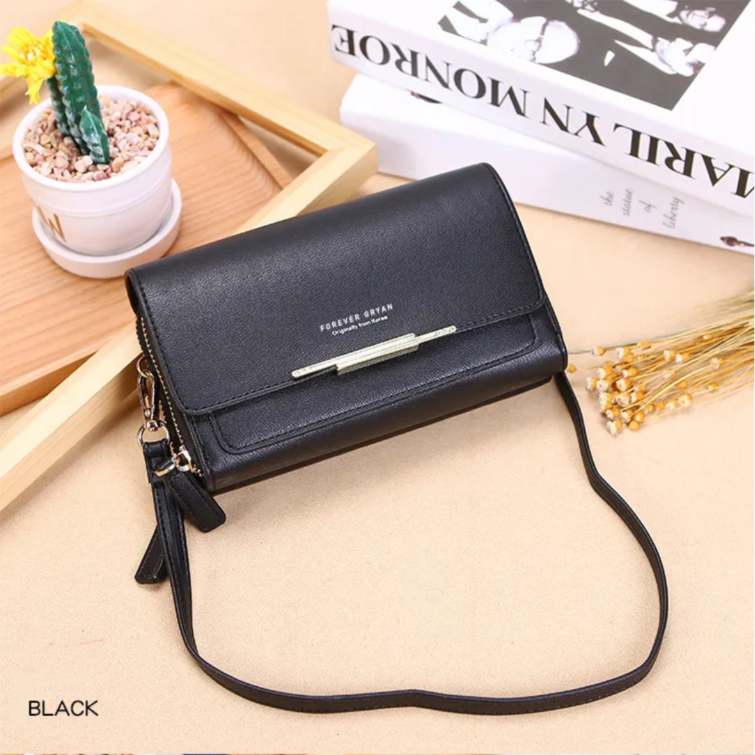 Black leather crossbody bag with a metal accent bar on a tan surface, featuring books and a small potted cactus.