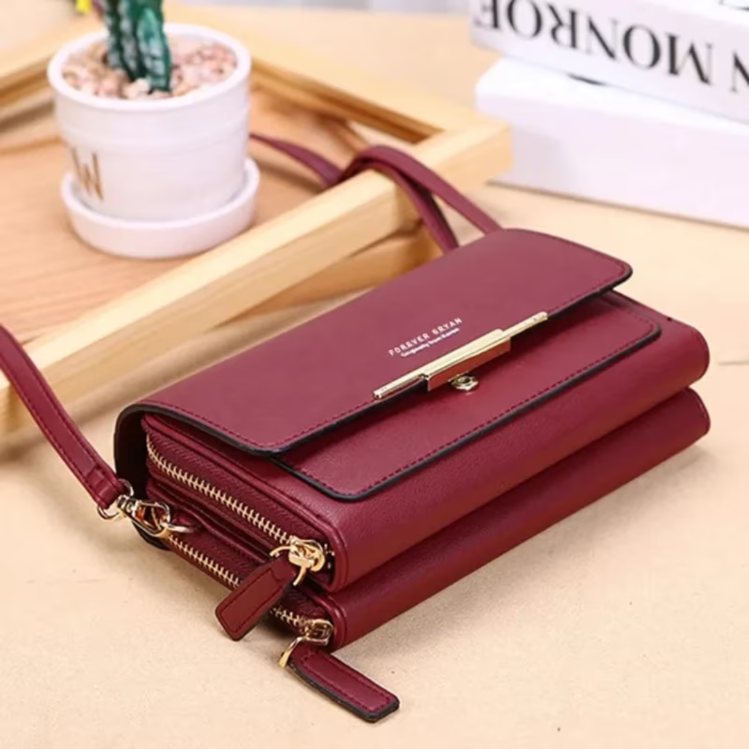 Maroon leather crossbody bag with dual zippered compartments, gold hardware, and shoulder strap on a desk.