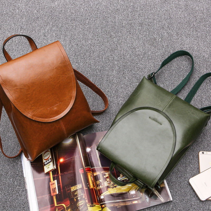 Fitlybuy tan and olive green leather backpacks with top flap closures on a gray background with a magazine and phone.