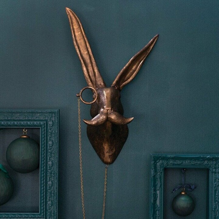 Bronze rabbit head wall decor with monocle and mustache on a dark teal wall with framed ornaments.