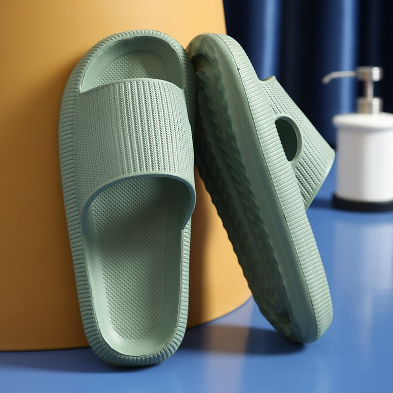 Sage green, textured EVA foam slide sandals leaning against a yellow backdrop with a soap dispenser in the background.