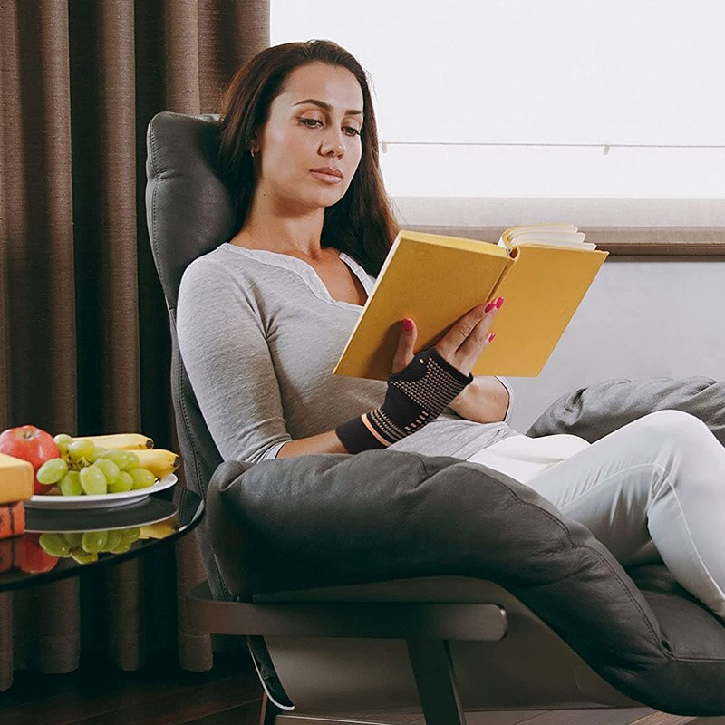Woman reading a book in a chair wearing a black and copper Fitlybuy compression wrist sleeve for heat therapy.