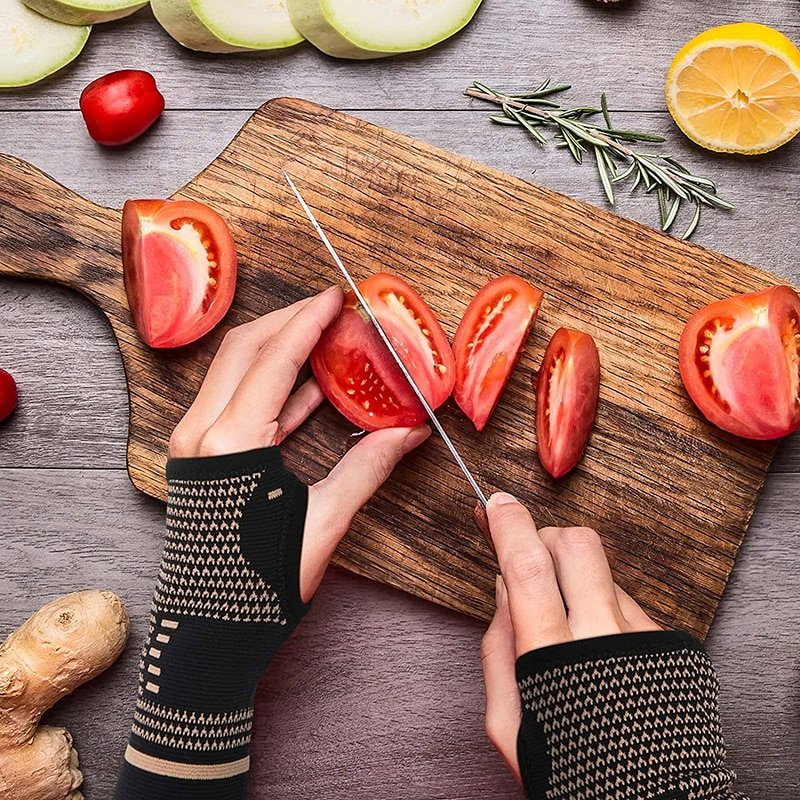 Hands wearing black and tan compression sleeves cutting tomatoes on a wooden board with produce.