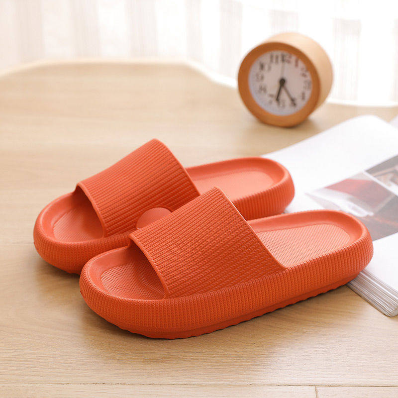Pair of bright orange Fitlybuy Cloud slippers with thick, textured soles on a wooden table near a clock and book.