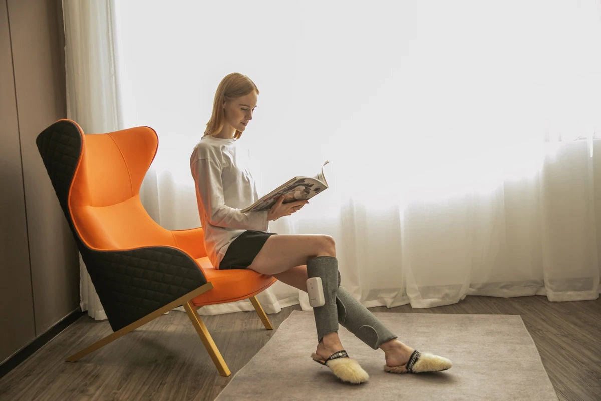 Woman reading in a chair wearing grey Fitlybuy air compression leg massagers for improved circulation.