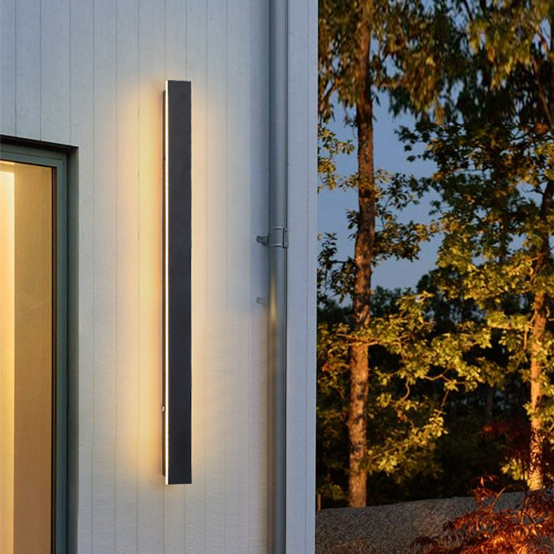 Minimalist black vertical LED outdoor wall light mounted on a white exterior wall at dusk.