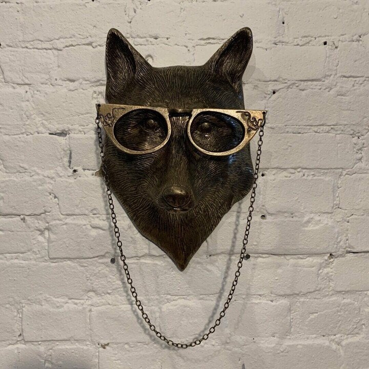 Bronze fox head wall decor featuring oversized gold-rimmed glasses with a dangling chain on a white brick background.