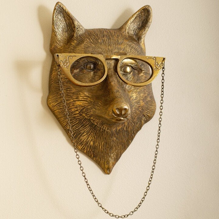 Gold-colored fox head wall decor featuring ornate cat-eye glasses with an attached hanging chain on a beige wall.