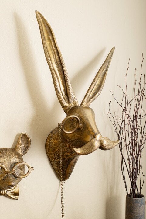 Gold-colored metal rabbit wall decor featuring a sculpted mustache and a monocle on a chain, mounted on a light wall.