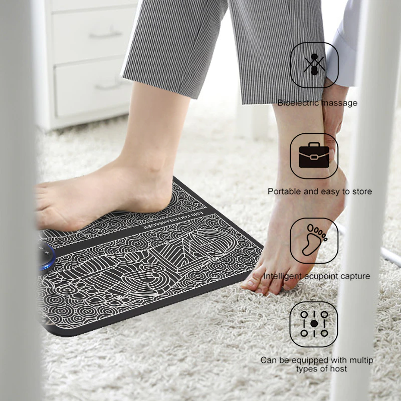 Black Fitlybuy EMS foot massager pad with white patterned surface, used under a desk to provide electric therapy.