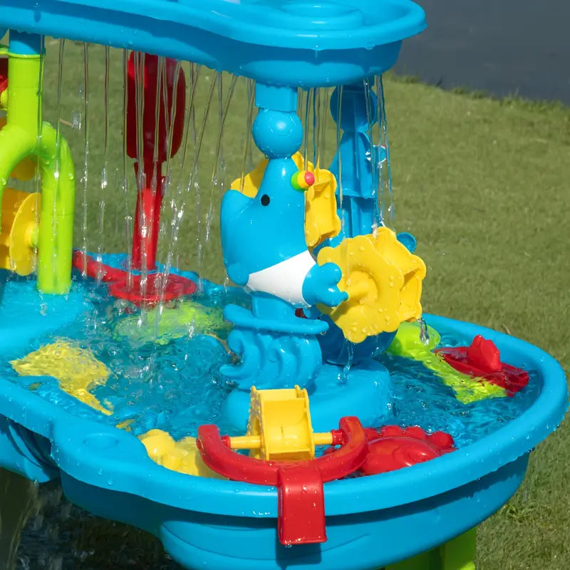 Fitlybuy 3-tier blue sand and water table with SplashGuard, fountain features, and colorful toys on a grass lawn.